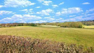 A wide-open grassy field under a bright blue sky with scattered white clouds. The landscape includes distant trees and rolling hills. A neatly trimmed hedge frames the bottom of the image. This plot of land, known as Km1 Underriver Sevenoaks, is periodically for sale with World of Auctions and Palace Auctions London.