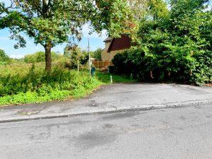 A narrow rural road bordered by greenery leads to a hidden residence partly obscured by dense trees and bushes. This unique land at Staines with future hope value for residential properties is up for sale by auction at World of Auctions. A small pole stands near the entrance, which has a visible driveway with some uneven and cracked pavement. A wooden fence is seen in the background.