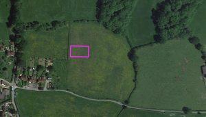 An aerial view of a landscape featuring green fields and dense tree areas. There is a small village with residential buildings to the left. In the center, a rectangular outlined plot of land stands prominently in the middle of the fields, periodically for sale with Palace Auctions London and World of Auctions.