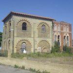 Property for sale by Auction An old, abandoned brick building with arched windows, partially deteriorated, and overgrown vines covering some sections. Standing on an unkempt lot with scattered wild plants under a clear blue sky, it evokes memories of Sheerness before the Water Tower development transformed the area. Presented by World of Auctions