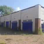 Property for sale by Auction A brick building with blue shutters on each of its three visible sections, surrounded by overgrown weeds and tall grass. The building appears to be abandoned, under a partly cloudy sky with a tree in the background. Nearby houses of The Water Tower development in Sheerness are also partially visible. Presented by World of Auctions