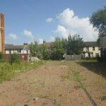 Property for sale by Auction A vacant, overgrown lot with patches of weeds and greenery. To the left, an old, partly dilapidated brick building, and to the right, a row of garages with blue doors. Residential houses and trees are visible in the background under a blue sky with scattered clouds near The Water Tower development in Sheerness. Presented by World of Auctions