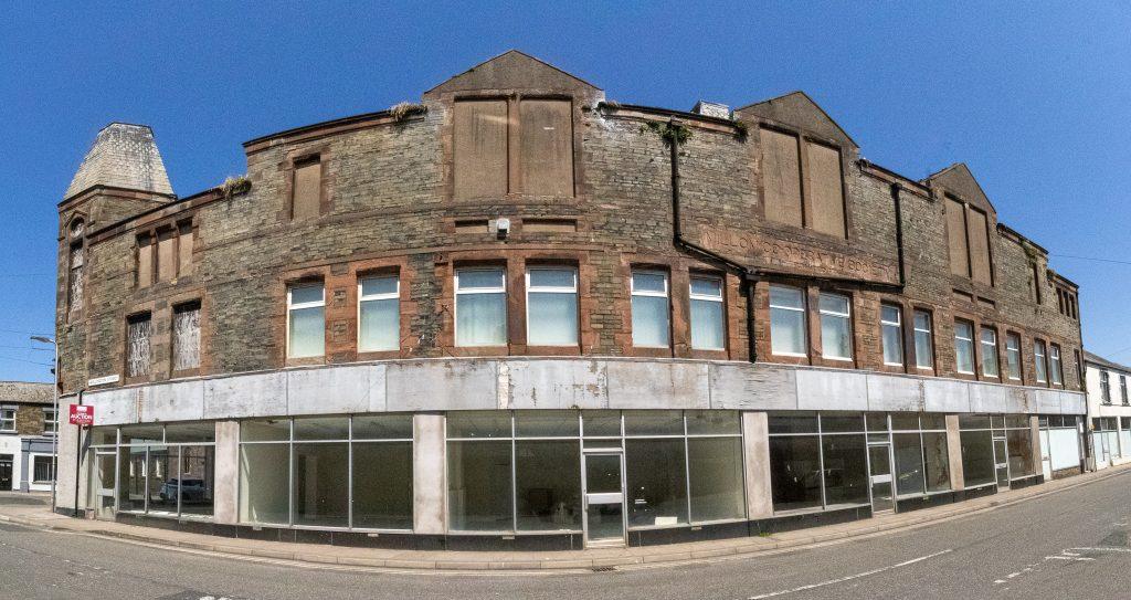 Property for sale by Auction A wide-angle view of a large abandoned building with boarded-up windows and dilapidated brickwork. The structure is on a street corner and has a row of empty storefronts with peeling paint and broken glass. The clear blue sky contrasts with the building's worn appearance. Presented by World of Auctions