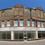 Property for sale by Auction A wide-angle view of a large abandoned building with boarded-up windows and dilapidated brickwork. The structure is on a street corner and has a row of empty storefronts with peeling paint and broken glass. The clear blue sky contrasts with the building's worn appearance. Presented by World of Auctions