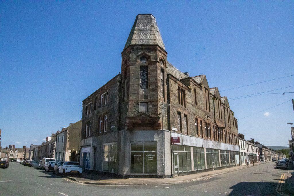 Property for sale by Auction A street corner featuring a large, historic, multi-story stone building with an angled, turreted roof. The building has large windows on the ground level, some covered with white shades. Parked cars line the street on a bright, sunny day under a clear blue sky. Presented by World of Auctions