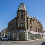 Property for sale by Auction A street corner featuring a large, historic, multi-story stone building with an angled, turreted roof. The building has large windows on the ground level, some covered with white shades. Parked cars line the street on a bright, sunny day under a clear blue sky. Presented by World of Auctions