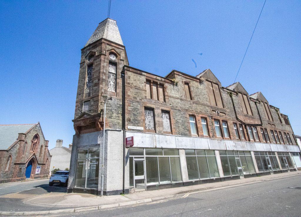 Property for sale by Auction An old, abandoned building made of stone with boarded-up windows stands under a clear, blue sky. The structure has a prominent tower on one corner and a row of large glass windows on the bottom floor. Next to it is another building with a peaked roof. Presented by World of Auctions