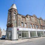 Property for sale by Auction An old, abandoned building made of stone with boarded-up windows stands under a clear, blue sky. The structure has a prominent tower on one corner and a row of large glass windows on the bottom floor. Next to it is another building with a peaked roof. Presented by World of Auctions