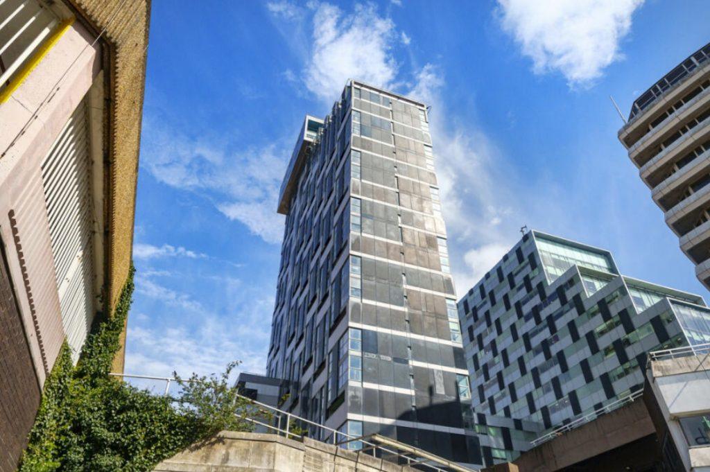 Property for sale by Auction View from the ground of three modern high-rise buildings with varying architectural styles against a bright blue sky with scattered clouds. One building has ivy climbing its wall, creating a lush facade. The scene, which could be home to a Liverpool penthouse investment apartment, includes a concrete structure in the foreground. Presented by World of Auctions