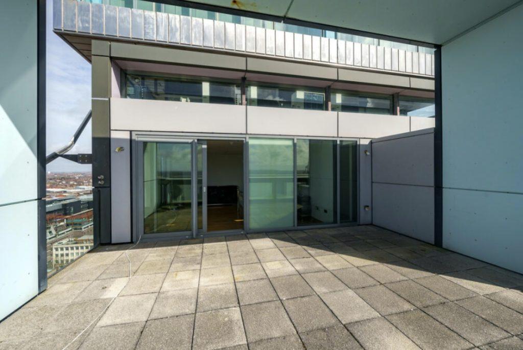 Property for sale by Auction A spacious balcony with a cement floor offers views of the city. Glass doors and windows lead to the interior of a modern Liverpool penthouse investment apartment. Metal and glass structures surround the area, providing a sleek and contemporary feel. The sky is clear in the background. Presented by World of Auctions