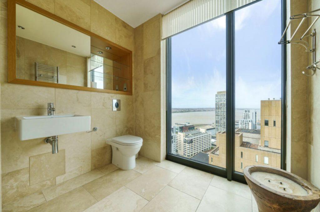 Property for sale by Auction A modern bathroom in a Liverpool penthouse investment apartment, featuring beige marble walls and floors, a floating sink, toilet, and large window showcasing cityscape views with buildings and a river. The room includes built-in shelves with a mirror and a rustic bowl-style basin. Presented by World of Auctions