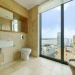 Property for sale by Auction A modern bathroom in a Liverpool penthouse investment apartment, featuring beige marble walls and floors, a floating sink, toilet, and large window showcasing cityscape views with buildings and a river. The room includes built-in shelves with a mirror and a rustic bowl-style basin. Presented by World of Auctions