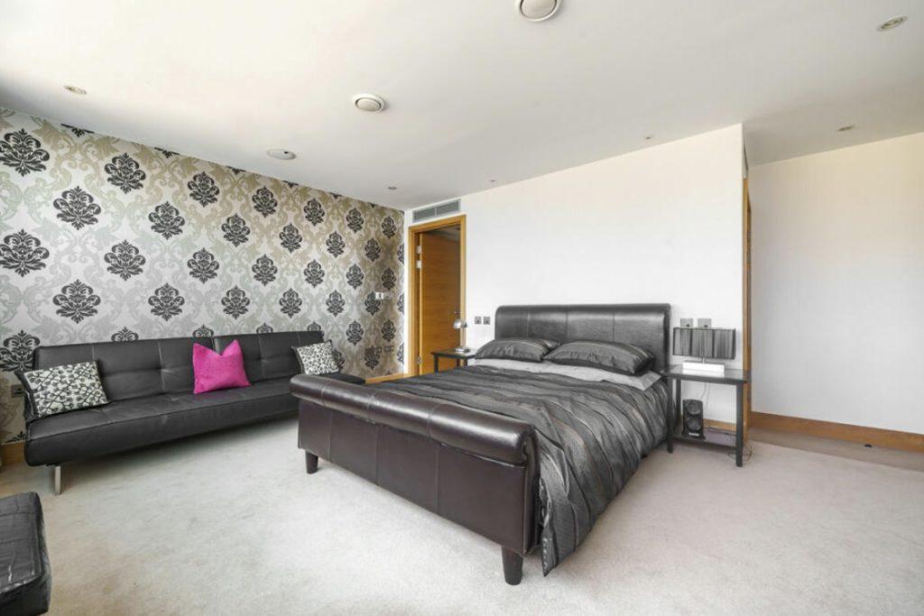 Property for sale by Auction A modern Liverpool penthouse investment apartment features a bedroom with a leather bed frame, grey bedding, and black nightstands. A black sofa with decorative pillows and a pink accent pillow sits against a wall adorned with black and silver damask wallpaper. The room boasts light carpeting and recessed lighting. Presented by World of Auctions