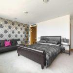 Property for sale by Auction A modern Liverpool penthouse investment apartment features a bedroom with a leather bed frame, grey bedding, and black nightstands. A black sofa with decorative pillows and a pink accent pillow sits against a wall adorned with black and silver damask wallpaper. The room boasts light carpeting and recessed lighting. Presented by World of Auctions