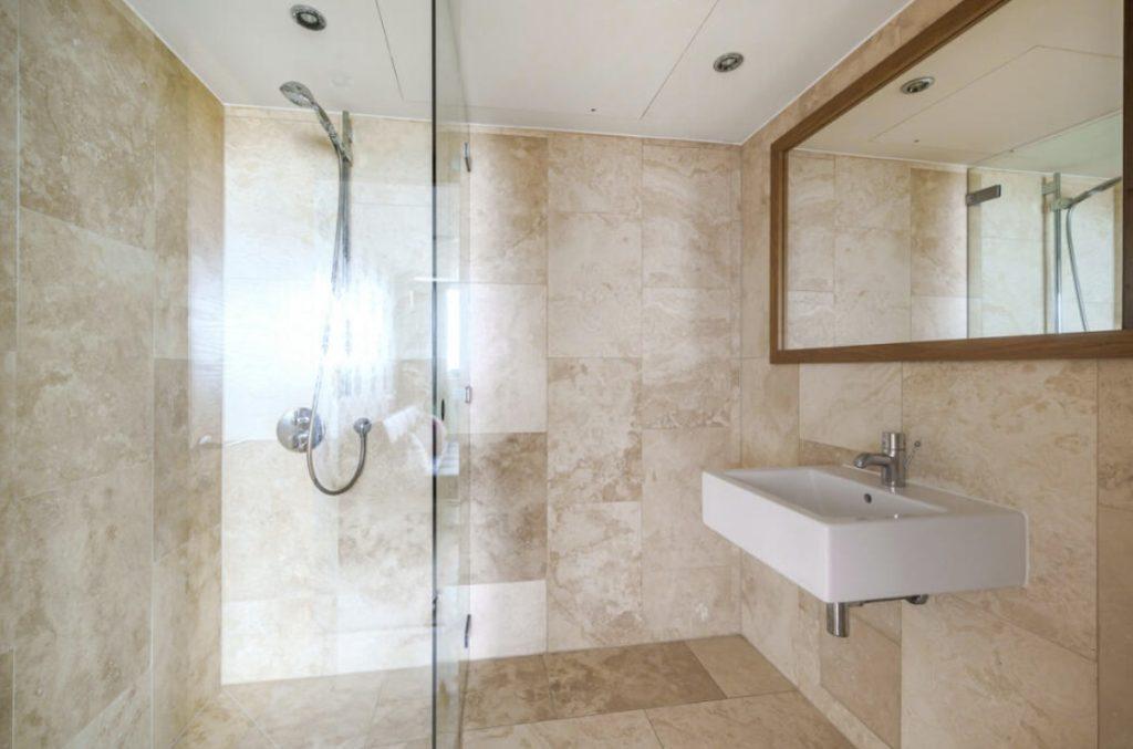 Property for sale by Auction A modern bathroom in a Liverpool penthouse investment apartment with beige tile walls and floors. It features a glass-enclosed shower with a wall-mounted shower head on the left. On the right, there is a rectangular white sink with a silver faucet and a large mirror above it. Presented by World of Auctions