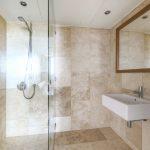 Property for sale by Auction A modern bathroom in a Liverpool penthouse investment apartment with beige tile walls and floors. It features a glass-enclosed shower with a wall-mounted shower head on the left. On the right, there is a rectangular white sink with a silver faucet and a large mirror above it. Presented by World of Auctions