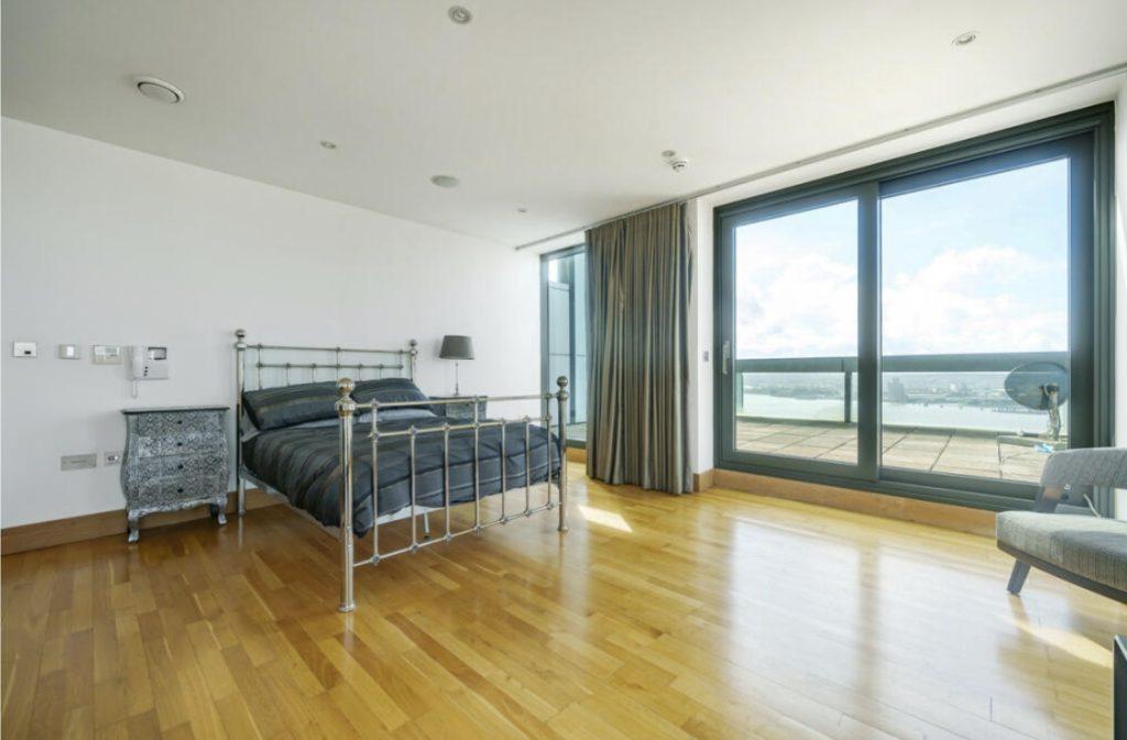 Property for sale by Auction A spacious, modern bedroom in a Liverpool penthouse investment apartment, featuring wooden flooring and a metal-frame bed with dark bedding. An ornate bedside table with a lamp and a large floor-to-ceiling window opens to a balcony with a scenic view. A gray chair is positioned near the window. Presented by World of Auctions