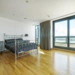 Property for sale by Auction A spacious, modern bedroom in a Liverpool penthouse investment apartment, featuring wooden flooring and a metal-frame bed with dark bedding. An ornate bedside table with a lamp and a large floor-to-ceiling window opens to a balcony with a scenic view. A gray chair is positioned near the window. Presented by World of Auctions
