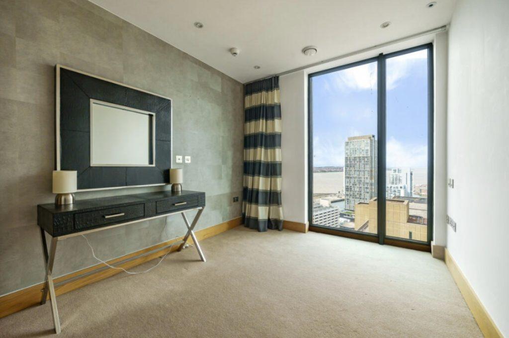 Property for sale by Auction A minimalist room in a Liverpool penthouse investment apartment boasts a large window offering city and river views. The room features a modern table with two lamps, a framed mirror on the wall, striped curtains, light wooden accents, and neutral-colored walls. Presented by World of Auctions