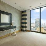 Property for sale by Auction A minimalist room in a Liverpool penthouse investment apartment boasts a large window offering city and river views. The room features a modern table with two lamps, a framed mirror on the wall, striped curtains, light wooden accents, and neutral-colored walls. Presented by World of Auctions