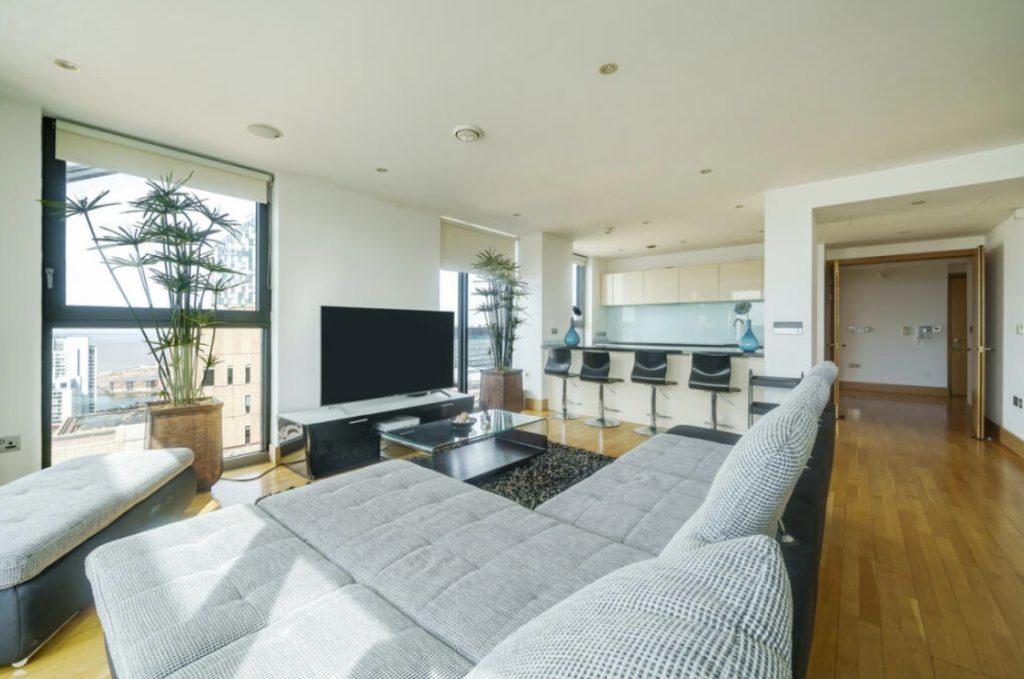 Property for sale by Auction A Liverpool penthouse investment apartment features a modern living room with light wood flooring, a large gray sectional sofa, and a flat-screen TV on a sleek black stand. Tall plants are beside the windows, and a kitchen with white cabinets and a breakfast bar with black high chairs is visible in the background. Presented by World of Auctions