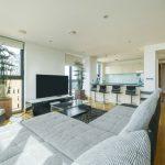 Property for sale by Auction A Liverpool penthouse investment apartment features a modern living room with light wood flooring, a large gray sectional sofa, and a flat-screen TV on a sleek black stand. Tall plants are beside the windows, and a kitchen with white cabinets and a breakfast bar with black high chairs is visible in the background. Presented by World of Auctions