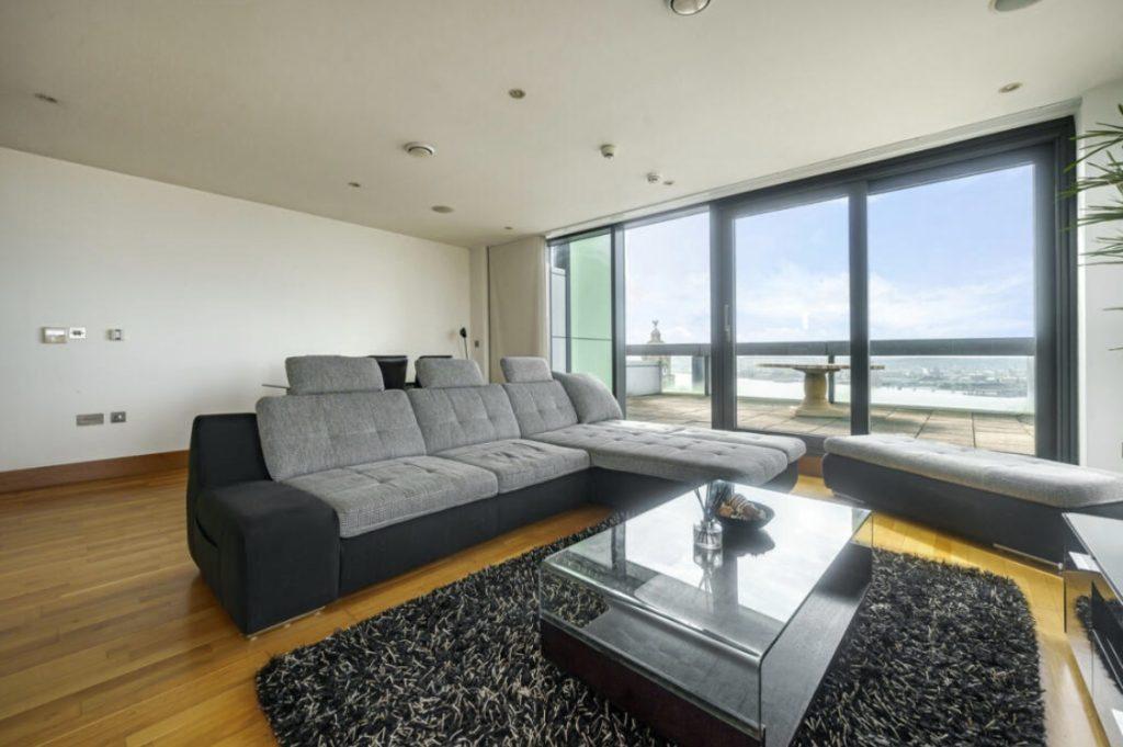 Property for sale by Auction A modern living room in a Liverpool penthouse investment apartment features a large gray sectional sofa and matching ottoman on a black shag rug. A glass-top coffee table sits in front of the sofa, while floor-to-ceiling windows and doors offer an expansive view of the balcony and cityscape beyond. Presented by World of Auctions