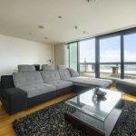 Property for sale by Auction A modern living room in a Liverpool penthouse investment apartment features a large gray sectional sofa and matching ottoman on a black shag rug. A glass-top coffee table sits in front of the sofa, while floor-to-ceiling windows and doors offer an expansive view of the balcony and cityscape beyond. Presented by World of Auctions