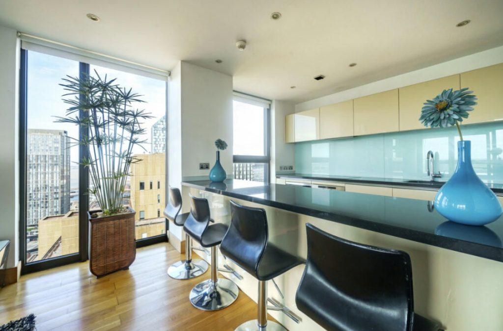 Property for sale by Auction Modern kitchen in a Liverpool penthouse investment apartment features a large island with a black countertop and four matching bar stools. Behind the island, light-colored cabinets, a built-in sink, and large windows provide natural light. Decor includes a tall plant and blue vases with flowers. Presented by World of Auctions