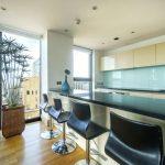 Property for sale by Auction Modern kitchen in a Liverpool penthouse investment apartment features a large island with a black countertop and four matching bar stools. Behind the island, light-colored cabinets, a built-in sink, and large windows provide natural light. Decor includes a tall plant and blue vases with flowers. Presented by World of Auctions