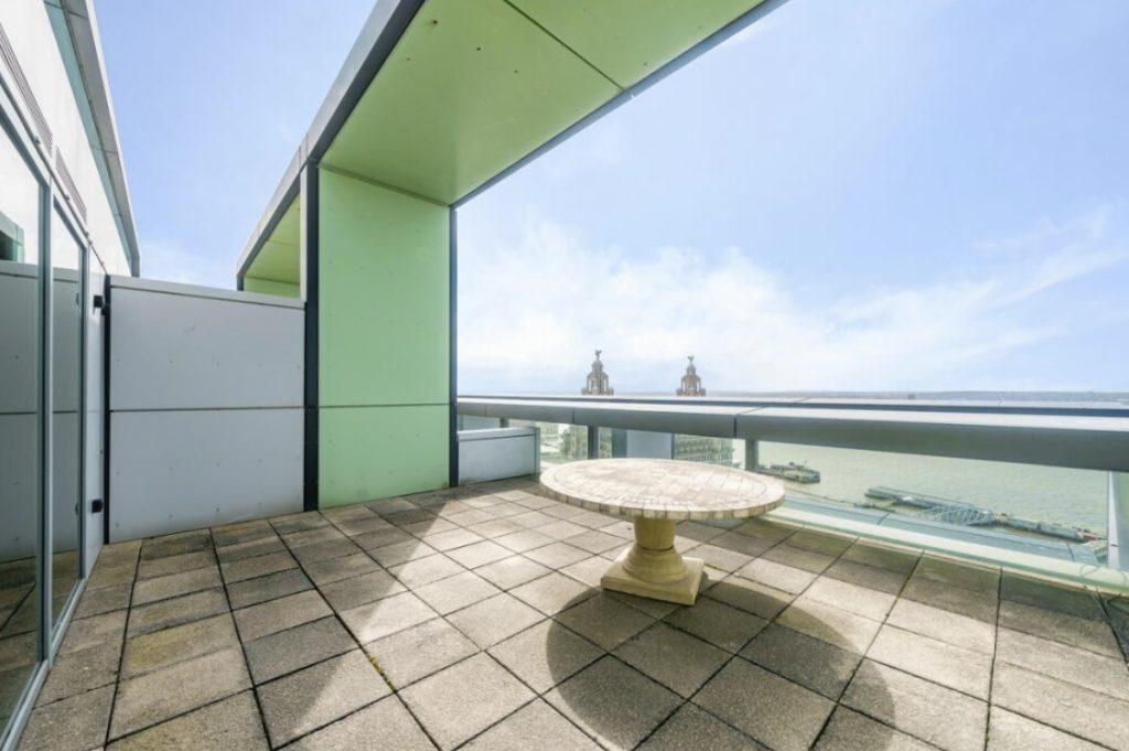 Property for sale by Auction A spacious rooftop terrace in a Liverpool penthouse investment apartment features a green and gray color scheme with a circular stone table at its center. The terrace offers a stunning view of the river and distant buildings under a clear blue sky. Presented by World of Auctions