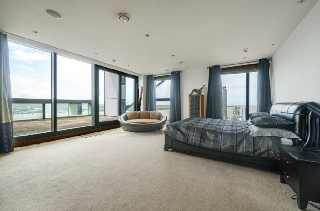 Property for sale by Auction A spacious bedroom in this Liverpool penthouse investment apartment features a large bed with dark bedding, a circular cushioned chair, a tall dresser, and floor-to-ceiling windows offering a panoramic city view. The room is decorated in neutral tones with blue accents and has a door leading to a private balcony. Presented by World of Auctions