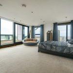 Property for sale by Auction A spacious bedroom in this Liverpool penthouse investment apartment features a large bed with dark bedding, a circular cushioned chair, a tall dresser, and floor-to-ceiling windows offering a panoramic city view. The room is decorated in neutral tones with blue accents and has a door leading to a private balcony. Presented by World of Auctions