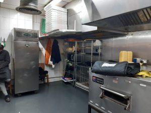 Property for sale by Auction A commercial kitchen with stainless steel appliances, shelves stocked with containers, and pizza boxes stacked high reflects the bustling activity of Pizza Shop 4 U. A person sits on a bench to the left, partially visible. An oven, refrigerator, and cooking hood are prominent in this mixed commercial and residential leasehold investment property. Presented by World of Auctions