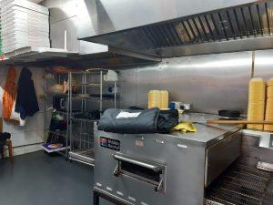 Property for sale by Auction A commercial kitchen in a bustling pizza shop 4 u, featuring a large industrial oven with a bag on top, shelving with pizza boxes, a microwave, and various supplies. The stainless steel walls and large ventilation hoods add to its efficiency. Yellow containers are stacked on a counter in the background. Presented by World of Auctions