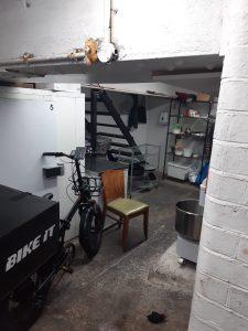 Property for sale by Auction A basement area with white brick walls and various items scattered around, reminiscent of a storage space in a mixed commercial and residential leasehold investment property. There's a bike labeled "BIKE IT" near a white cabinet, a wooden chair with a green seat in the foreground, metal shelves, and a staircase leading to an upper level. Presented by World of Auctions