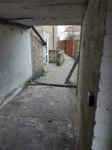 Property for sale by Auction A narrow, semi-enclosed passageway with bare concrete and brick walls leads to an open, paved area. The ground appears old and worn, with scattered debris. In the distance, there's a brick building housing a pizza shop 4 u alongside other mixed commercial and residential leasehold investment properties. Presented by World of Auctions