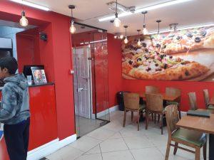 Property for sale by Auction A small pizza shop 4 u with bright red walls, wooden chairs, and tables. The wall features a large, vibrant image of pizzas. Edison bulbs hang from the ceiling. A boy in a hoodie stands near the counter on the left. The décor is clean and modern, making it an attractive mixed commercial and residential leasehold investment property. Presented by World of Auctions