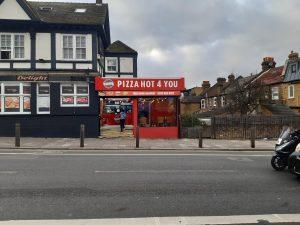 Property for sale by Auction A street view of "Pizza Hot 4 You," a pizza shop with a red and white storefront in a mixed commercial and residential area. The building, featuring black trim, is next to homes, making it an attractive leasehold investment property. A man on a motorcycle rides past the shop on the road. Presented by World of Auctions