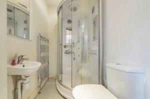 Property for sale by Auction A small bathroom on Rowan Avenue features a white sink on the left with a wall-mounted mirror above it, a silver towel radiator on the left wall, a rounded glass shower enclosure in the center, and a white toilet on the right side against a pale-colored wall. Presented by World of Auctions