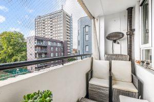 A small balcony at Flat 3, 1 Macclesfield House, Central Street, LONDON EC1V 8BA features a wicker chair with beige cushions and is surrounded by a safety net. Potted plants grace the table in the foreground as sunlight filters in, illuminating nearby buildings, trees, and a blue sky. Ideal investment for sale by auction. Presented by World of Auctions
