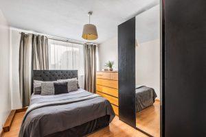 Modern bedroom featuring a neatly made bed with grey bedding, framed by a tufted headboard. A wooden dresser with a potted plant atop it stands against the wall, opposite a large mirrored wardrobe. Curtains cover the window, and a ceiling light hangs above. Flat 3,1 Macclesfield House Central Street LONDON EC1V 8BA investment for sale by auction. Presented by World of Auctions