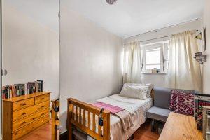 Flat 3 at Macclesfield House, Central Street, LONDON EC1V 8BA features a small, cozy bedroom with a single bed adorned with a white and pink blanket. Beside the bed sits a grey chair and a light wooden chest of drawers topped with books. Natural light filters through the window with white curtains, highlighting the room's light-colored walls and hardwood flooring. This investment Presented by World of Auctions