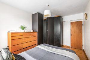 A cozy bedroom in Flat 3, 1 Macclesfield House, Central Street LONDON EC1V 8BA features a large wooden dresser with a potted plant and books on top. The bed is neatly made with grey and white bedding. Black wardrobes occupy a corner, and a wooden door leads out of the room. A white ceiling light fixture hangs above. Presented by World of Auctions