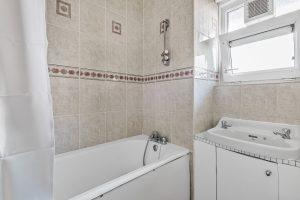 A bathroom in Flat 3 with beige tiled walls and a white bathtub. Above the bathtub is a showerhead and faucets, with a curtain on one side. A small sink with a cabinet sits below an open window, making it an attractive feature of Macclesfield House Central Street LONDON EC1V 8BA investment for sale by auction. Presented by World of Auctions