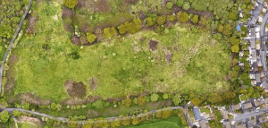 Property for sale by Auction Aerial view of a large open green field with patches of trees and shrubs along the perimeter. A curved road runs along the bottom and right edges of the image. Houses are clustered in the bottom right corner near the road. Presented by World of Auctions