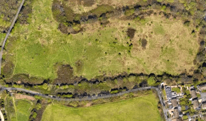 Property for sale by Auction Aerial view of a landscape featuring a large, open grassy area with patches of trees and shrubs. A narrow road runs diagonally near the bottom, with a few buildings and houses clustered on the right side. Pathways meander through the greenery on the left. Presented by World of Auctions