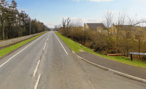 Property for sale by Auction A rural road stretches into the horizon, flanked by grassy areas and leafless trees. To the right, a sidewalk leads to several houses, and a sign indicates the direction towards Northampton. The sky overhead is partly cloudy, with patches of blue visible. Presented by World of Auctions