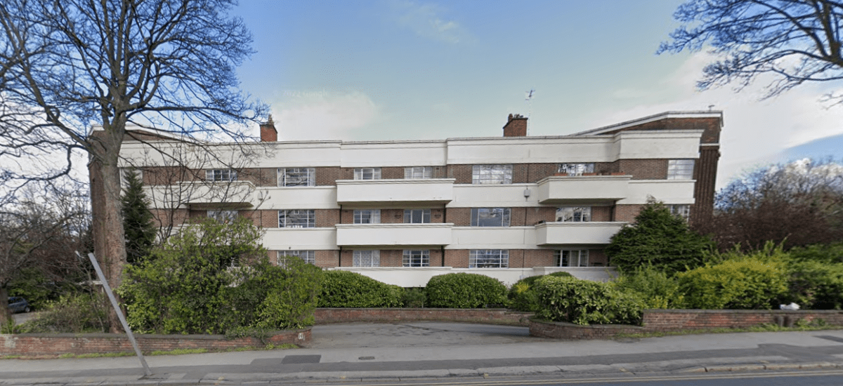 The three-story residential building at 29 Mansfield Court, Nottingham, features a mix of red brick and white concrete facade. Each apartment has a balcony, surrounded by trees and shrubbery. A paved driveway and sidewalk are visible in the foreground with a blue sky above. NG5 2BW for sale by auction with World of Auctions.