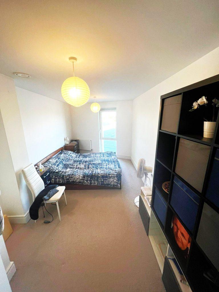 A cozy bedroom in a beautiful modern London apartment features a bed with a blue and white patterned duvet, a white chair with a black jacket draped over it, a black shelving unit, and a tall white cabinet. A glowing pendant lamp hangs from the ceiling, while the window with balcony door illuminates the space. This stunning property is available for sale by auction with World of Auctions and Palace Auctions London
